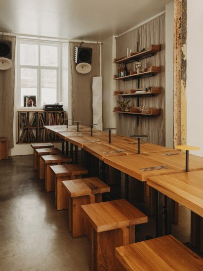 Photograph of a communal dining area made from small oakwood tabletops and matching geometric stools. Beneath a window at the rear of the dining area is a display cabinet exhibiting a large vinyl collection.