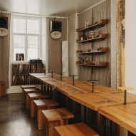 Photograph of a communal dining area made from small oakwood tabletops and matching geometric stools. Beneath a window at the rear of the dining area is a display cabinet exhibiting a large vinyl collection.