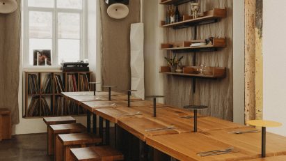 Photograph of a communal dining area made from small oakwood tabletops and matching geometric stools. Beneath a window at the rear of the dining area is a display cabinet exhibiting a large vinyl collection.