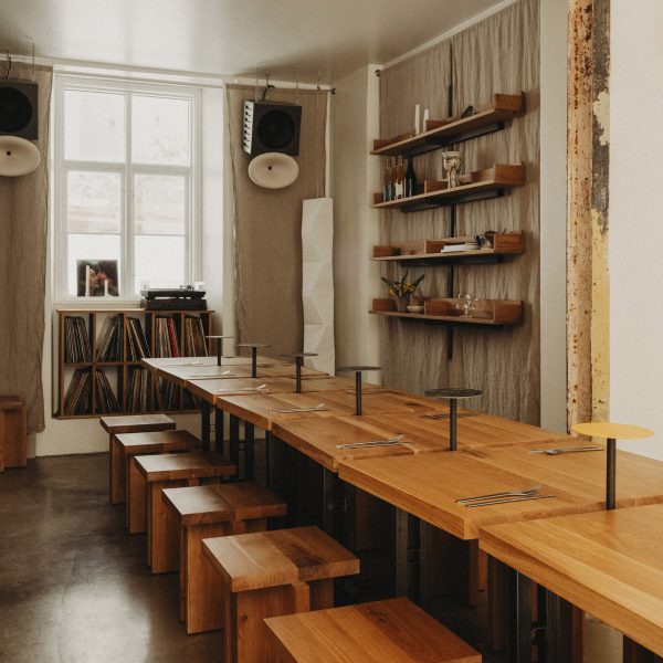 Photograph of a communal dining area made from small oakwood tabletops and matching geometric stools. Beneath a window at the rear of the dining area is a display cabinet exhibiting a large vinyl collection.