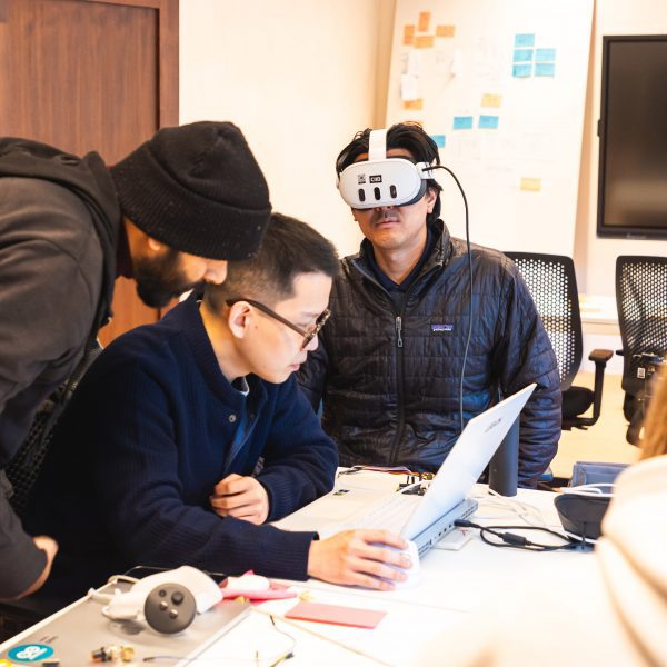 A photograph of students on an interaction design course. One is wearing a VR headset and the others are working at a laptop.