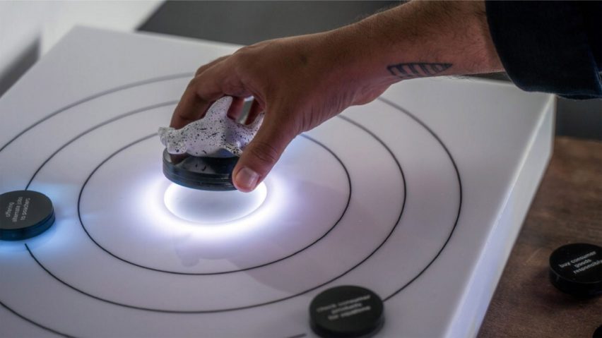 A photograph of a person's hand holding a rock over a white surface with black circles on it.