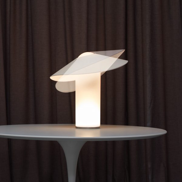 A photograph of a lamp in tones of white placed on a grey table with a brown backdrop