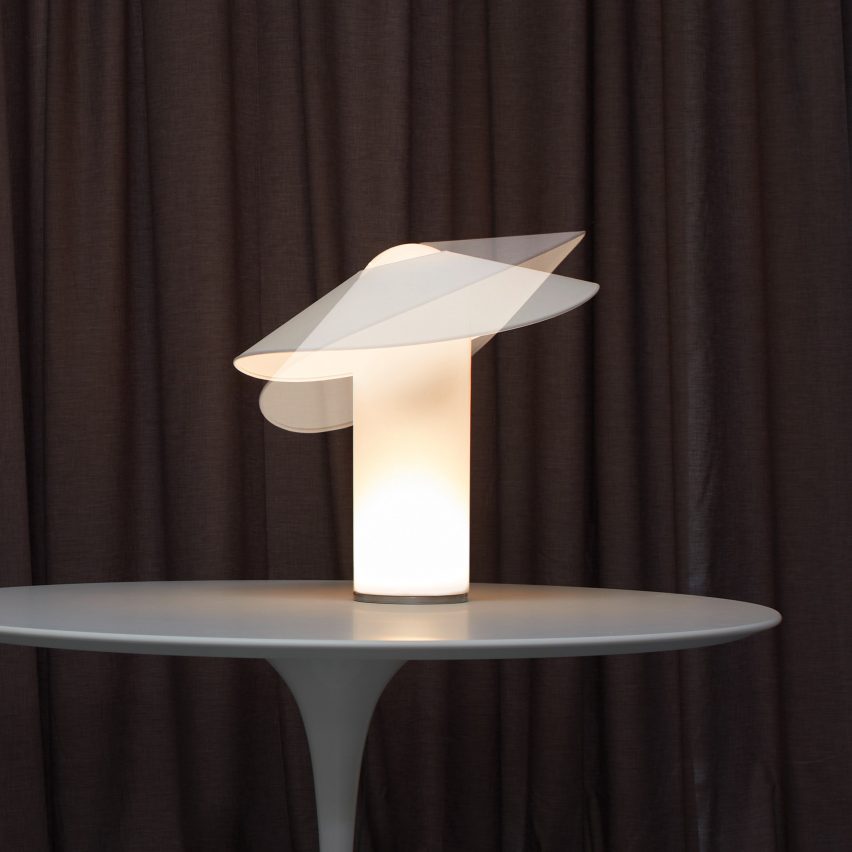 A photograph of a lamp in tones of white placed on a grey table with a brown backdrop