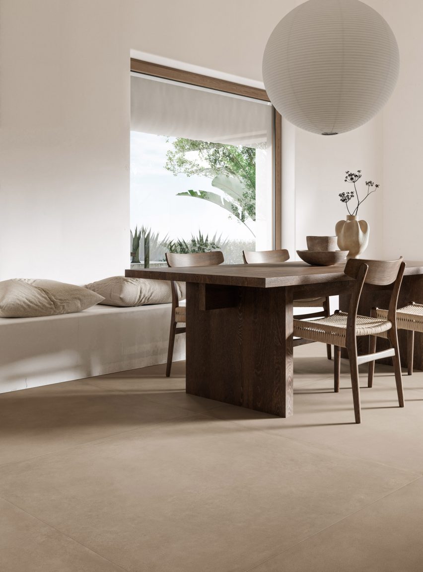 A photograph of an interior space with a brown wooden table and chairs and white seating area with white cushions. The floor is comprised of beige coloured tiles