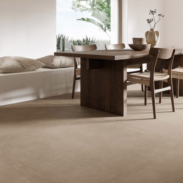 A photograph of an interior space with a brown wooden table and chairs and white seating area with white cushions. The floor is comprised of beige coloured tiles