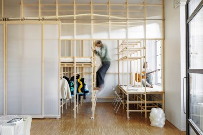 Photo of a bedroom built into a larger space using a fir wood structure and polycarbonate panels, showing a raised tatami sleeping platform on the left side with wardrobe storage underneath and on the right looking out of the window, another platform with chairs that functions as two desks
