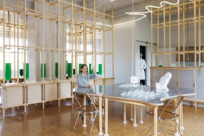 Photo of Claudia Pasquero sitting at a timber table with an unusually, wavy-shaped tabletop edge in front of a wall of photobeoreactors containing green liquid