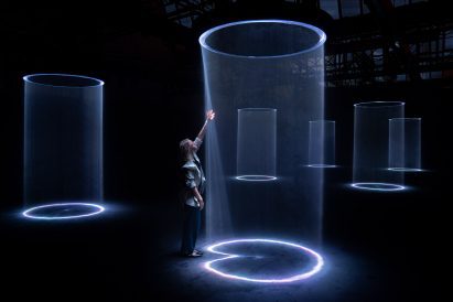 Photo of a person standing on the edge of a column of light within an artwork and reaching up to touch it