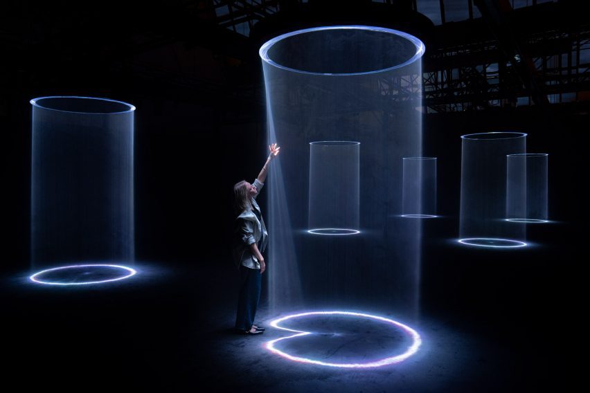 Photo of a person standing on the edge of a column of light within an artwork and reaching up to touch it