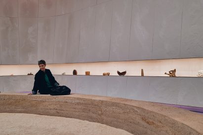 Photo of an exhibition space showing a woman sitting on a rammed earth bench seat that curves around, with sand in front and a display of objects along one linear shelf behind her