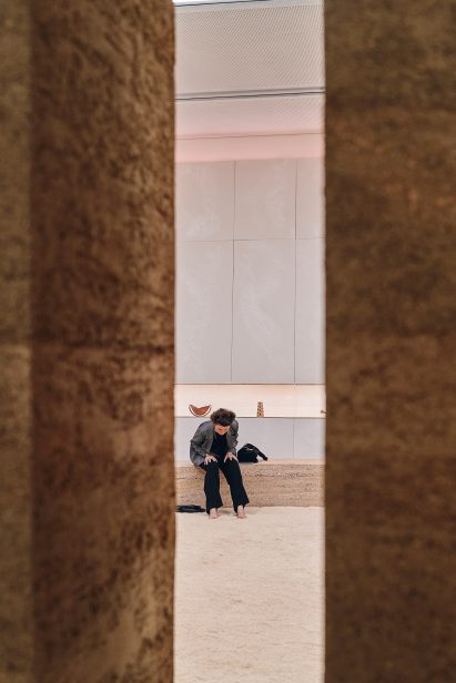 Photo looking through a clean-lined gap in a rammed-earth strcuture at a woman as she sits on bench seating and digs her bare feet into sand
