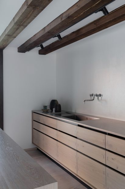 Wooden kitchen units beneath wall-mounted metal tap in a room with wooden ceiling beams