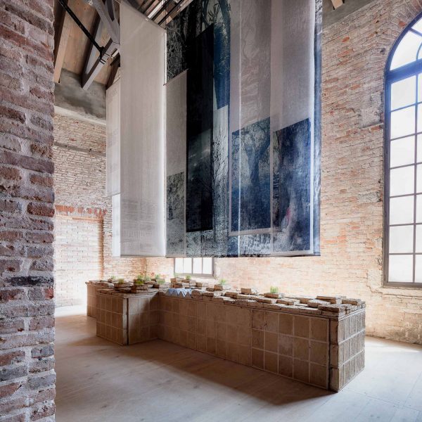 Photo of the Lebanese pavilion at the Venice Architecture Biennale 2025, showing a display table made of earthen bricks with textile panels hanging overhead