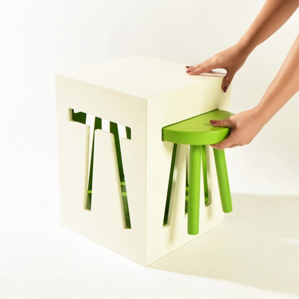 a photograph of a person interacting with an abstract stool design, featuring a white cube with a pull-out green stool in it.