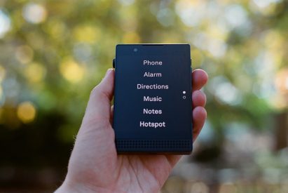 Photo of a person's hand holding up the Light Phone 3, which is open to its plain-text black-and-white menu reading phone, alarm, directions, music, notes and hotspot