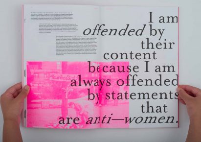 a photograph of a person holding an open book; pages showing black text and tones of white and pink