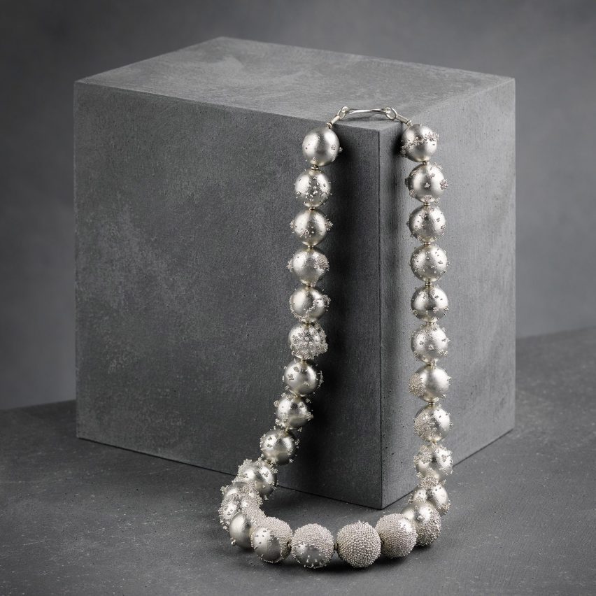 a photograph of a silver beaded necklace hanging from a corner of a grey cube
