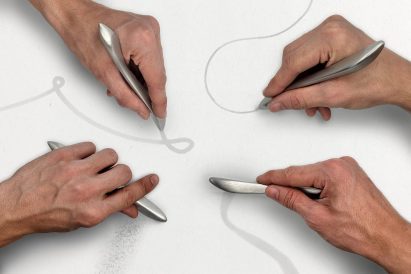 Four hands each holding silver coloured writing implements and making pale grey marks on a white surface with them