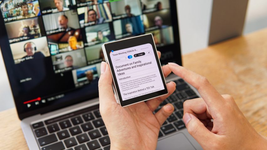 Photo of a person's hands holding up a small, square device with a document on it reading Document on Family Adventures and Inspirational Ideas. In the background their laptop screen shows a video call with many participants