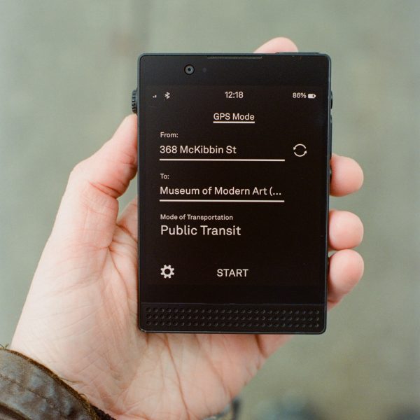 Photo of a person's hand holding a Light Phone 3 showing the directions app, with white text on a white background reading 'From 368 McKibben Street' 'To Museum of Modern Art' and a mode of transportation field set to 'public transit'. Below that a text button reads 'start'