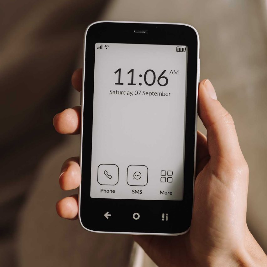 Photo of a hand holding the Mudita Kompakt showing a white e-ink screen showing the time and date at the top and three app icons along the bottom