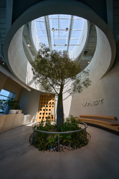 Photo of the lobby of Vast's headquarters in Long Beach, showing a room with chamfered corners, a tall tree planted in the ground, a circular oculus cut into the mezzanine above and a circular skylight beyond that