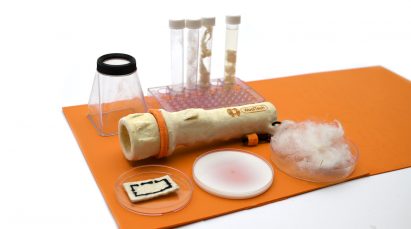Photo of the WoolTech torch laid on an orange mat and surrounded by material samples in test tubes and petri dishes