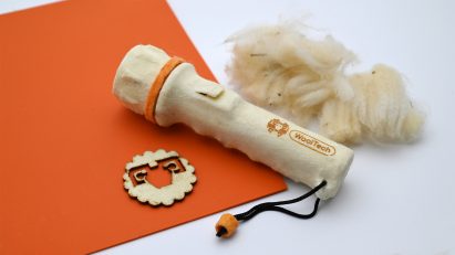 Photo of a torch made of a pale biomaterial with WoolTech branding on its casing, flatlayed on a surface with an orange card and a fluffy sample of wool