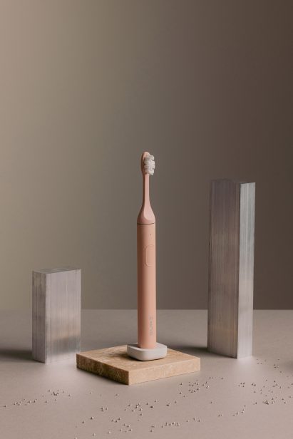 Studio photo of a pale pink Suri toothbrush on its small pebble-like charging stand sat among metal and stone plinths
