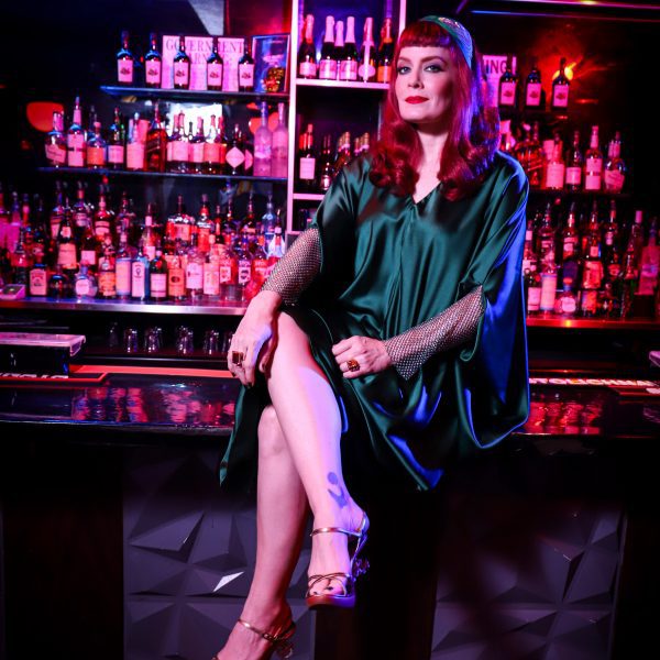 Ana Matronic at a bar counter in a dimly lit venue, wearing a flowing green satin dress and gold heels, ahead of starring at the Dezeen Awards 2025 party