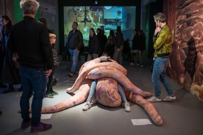 Photo of a person wearing VR goggles while they sit in a pale pink beanbag-like creation that wraps the sitter in its long limbs. The installation is within an exhibition environment and other visitors walk past looking on curiously