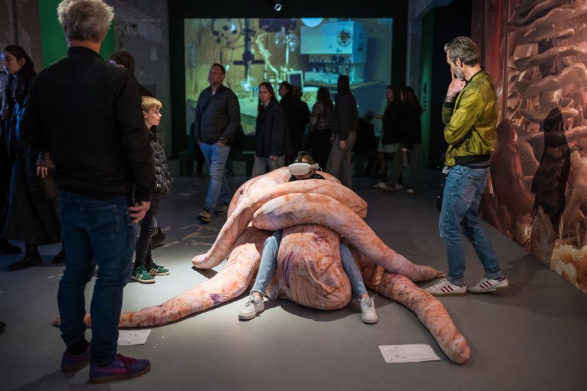 Photo of a person wearing VR goggles while they sit in a pale pink beanbag-like creation that wraps the sitter in its long limbs. The installation is within an exhibition environment and other visitors walk past looking on curiously