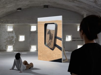 Photo of the Who Stole the Sky video installation by Ze Qiu showing a portrait-orientated large screen with an image on it of a horse traversing a desert landscape as seen through a car's side mirror. People are watching the work