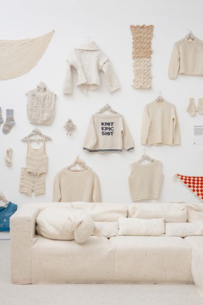 Wall of knitted garments in natural shades including a jumper that reads 'knit epic shit' at the Wool Rediscovered exhibition