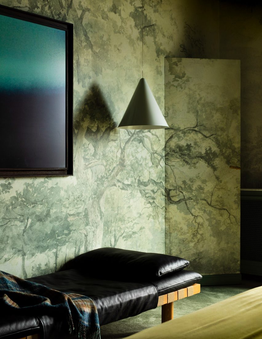 Photograph showing a green nature-inspired wallpaper, and a hanging pendant lamp over a wooden bench with a black leather covering.