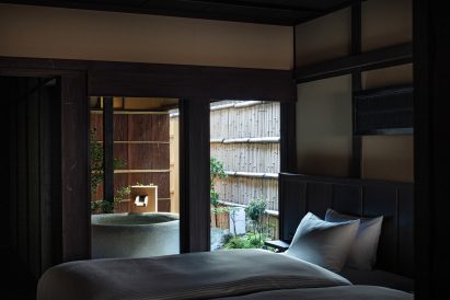 A photograph of a single bed positioned next door to two floor-to-ceiling glass windows that frame a large open-air bathtub.