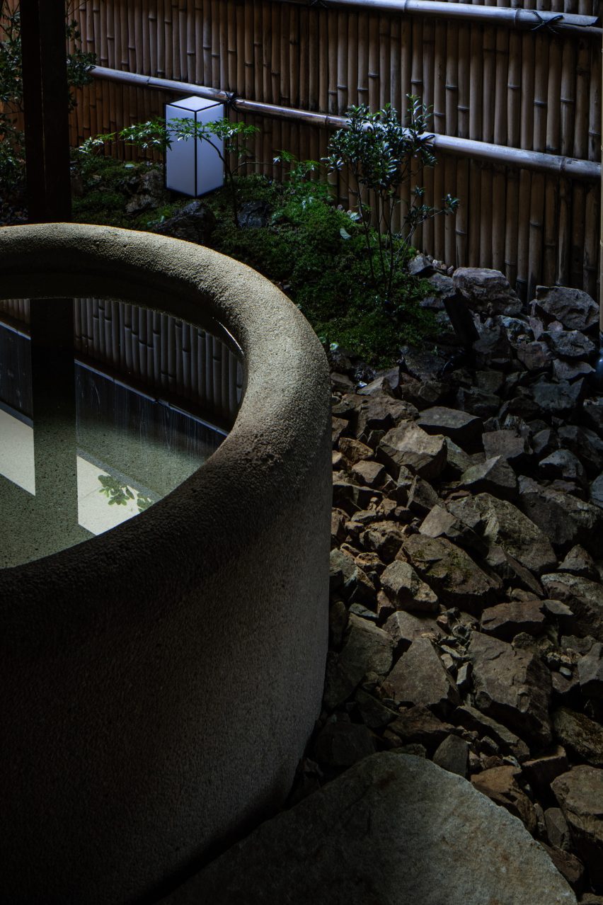 A close-up photograph of a circular, stone bathtub in an outdoor courtyard garden which features plants and a bamboo wall.