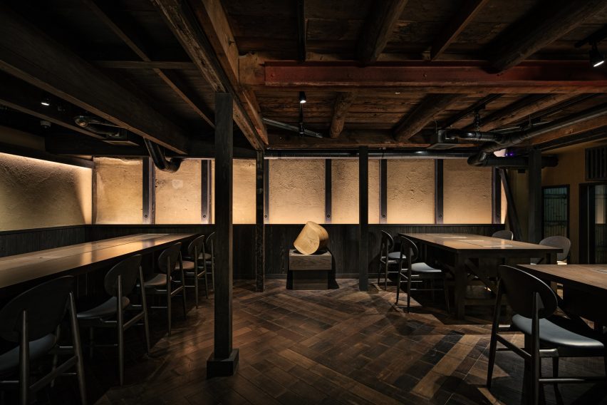 A warmly lit dining area with a dark wooden floor, ceiling and dining tables. A long dining table takes up the left side of the photo. Two shorter dining tables make up the right side of the photo. A small sculpture is in the back, centre of the image.