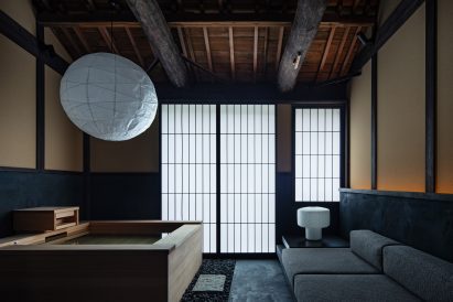 A wooden bath tub sits on the left side of the photo, with a large, white, paper lampshade hanging above it. The right side of the image shows a low, grey sofa facing the bathtub.