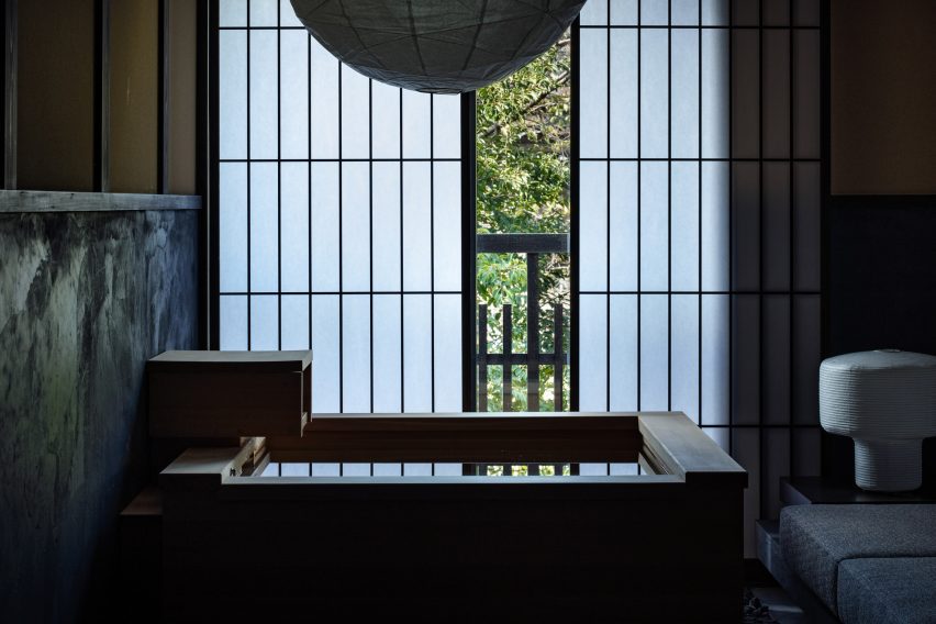 A photo of a large, square, wooden bathtub and behind it, latticed sliding doors made from washi-paper are slightly open to show the outdoor greenery.