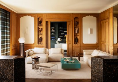 A picture of the large living room, showing the restored wooden panelling, a central seating area made up of modular white sofas and a clear acrylic feature-stool. There is a low, teal coffee table in a glossy finish. The back of the room shows an open walkway into the library.