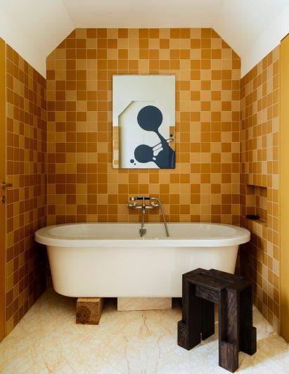 A picture of a bath which is tucked into a small space at the end of the bathroom. The bathroom displays monochromatic orange tiled walls. In the middle of the wall above the bath tub is a small abstract painting. The floor is white with an orange marbling effect. A small, black, wooden stool sits next to the bath tub to the right of the image.