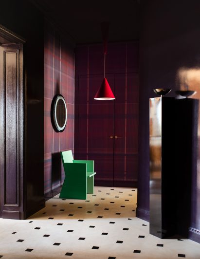 Photograph of the dramatic entrance hall, which has tartan wallcoverings in a deep purple. There is a patterned marble floor, and a green sculptural chair, spotlighted by a red-hanging pendant light.
