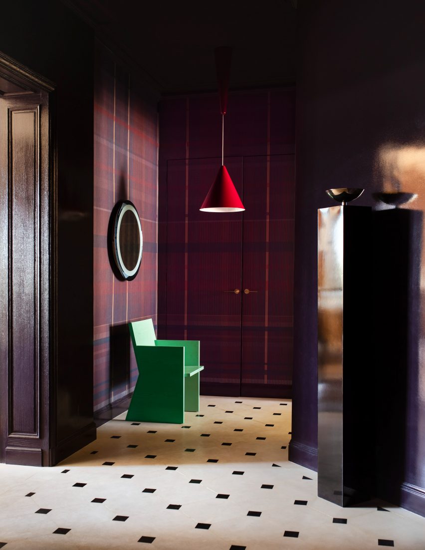 Photograph of the dramatic entrance hall, which has tartan wallcoverings in a deep purple. There is a patterned marble floor, and a green sculptural chair, spotlighted by a red-hanging pendant light.