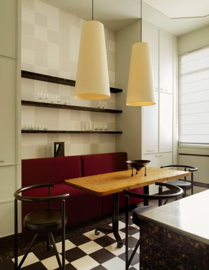 Photograph of the bold kitchen, showing a small seating area with red-velvet banquette seating and stylised chairs. The kitchen has a black and white checkerboard floor.