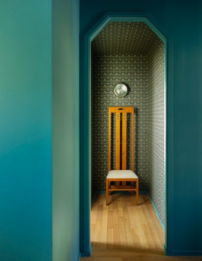 A picture taken from one side of a teal door frame, showing a wooden chair with a tall back rest. The floor is made from light wooden floorboards, and the wallpaper is dark green and intricately patterned.