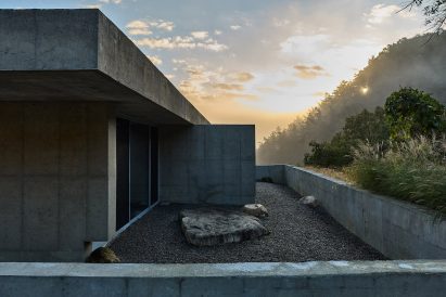 Concrete building at sunrise, with misty forested hills in the background and a gravel courtyard featuring large rocks in foreground