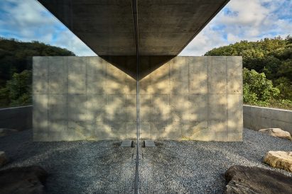 Mirrored glass surface reflecting concrete wall and gravel courtyard, with sunlight casting patterned shadows across the scene