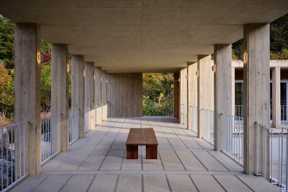 Covered concrete walkway with evenly spaced columns, featuring a wooden bench in the centre